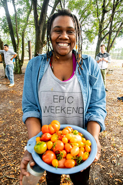 Smiling black woman holds a bowl of orange and yellow tomatoes towards the camera.